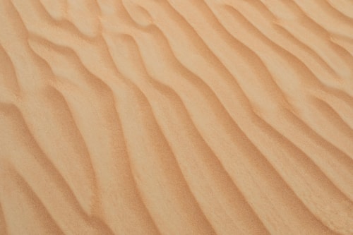 Gros plan sur des vagues de sable formées par le vent sur une plage, montrant une texture ondulée et des nuances de beige.