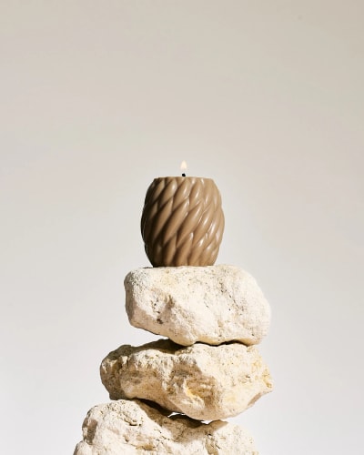 Bougie cube orange Fiamme composée de petites sphères, posée sur un miroir rond, avec un diffuseur d'arômes en verre brun et des bâtonnets en arrière-plan, le tout sur une surface claire.