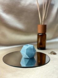 Bougie bleue géométrique Fiamme posée sur un miroir rond avec une bouteille en verre ambré contenant des bâtonnets de parfum en arrière-plan