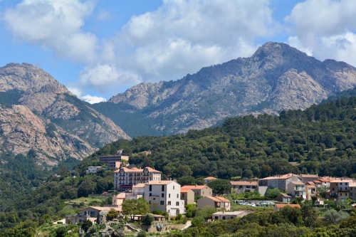 Village Corse ( Moca-Croce) avec maisons aux toits rouges entouré de collines boisées et montagnes rocheuses sous un ciel partiellement nuageux.