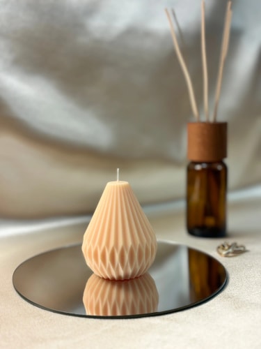 Bougie beige Fiamme en forme de goutte avec motif géométrique, posée sur un miroir rond, avec une bouteille en verre ambré et des bâtonnets de parfum en arrière-plan sur une surface beige