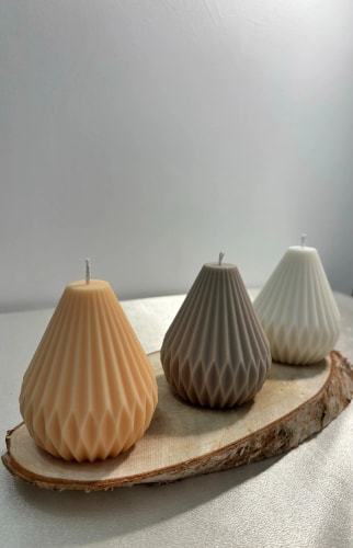 Trois bougies Fiamme décoratives de formes géométriques posées sur une tranche de bois, chacune d'une couleur différente : beige, marron clair et blanc.