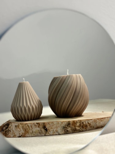 Deux bougies décoratives Fiamme de couleur marrons avec des motifs en relief, posées sur une tranche de bois, reflétées dans un miroir rond.