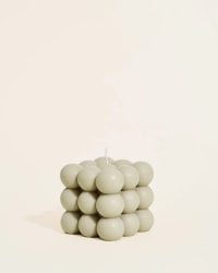 Bougie blanche Moca en forme de cube composée de petites sphères empilées, posée sur un miroir rond, avec un diffuseur en verre brun et des bâtonnets en arrière-plan sur une surface beige