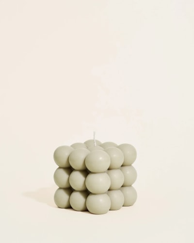 Bougie blanche Moca en forme de cube composée de petites sphères empilées, posée sur un miroir rond, avec un diffuseur en verre brun et des bâtonnets en arrière-plan sur une surface beige