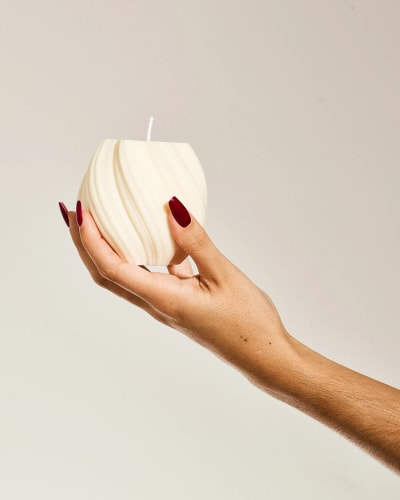 Bougie sculptée grise Fiamme posée sur un miroir rond avec un flacon brun et un anneau doré en arrière-plan sur un tissu beige.