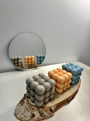 Bougies cube Fiamme composées de sphères de couleur grise, pêche et bleue, posées sur une tranche de bois, avec un miroir rond en arrière-plan reflétant les bougies.