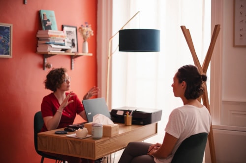 Une femme en haut rouge est assise à son bureau et discute avec une autre personne assise en face d'elle dans un cabinet aux murs orange.