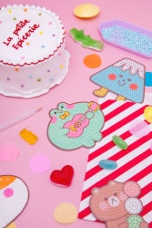 Composition de pâtisseries colorées sur fond rose : gâteau décoré « La Petite Épicerie », biscuit nuage fleuri, tablier, souris et étoile en sucre. Une nappe rayée rouge et blanche complète la scène.