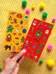 Deux mains tiennent une pochette colorée couverte d’illustrations et de motifs variés sur le thème du voyage au Japon, sur un fond orange et rose.