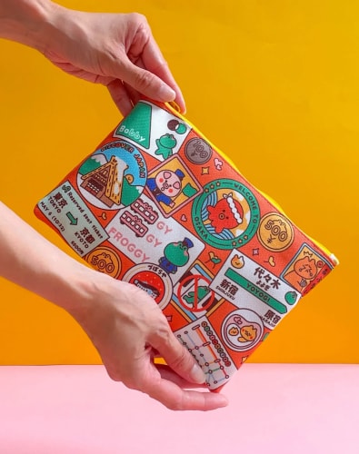 Deux mains tiennent une pochette colorée couverte d’illustrations et de motifs variés sur le thème du voyage au Japon, sur un fond orange et rose.