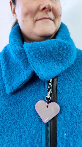 Gros plan d'un enfant portant une veste rose avec un réflecteur en forme de cœur accroché à la fermeture éclair. L'enfant s'appuie sur la main de son parent, l'atmosphère de l'image est chaleureuse et bienveillante.