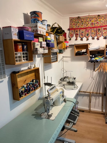 Petit atelier de couture encombré, avec machines à coudre, étagères de fournitures, tissus suspendus et décoration colorée au mur.