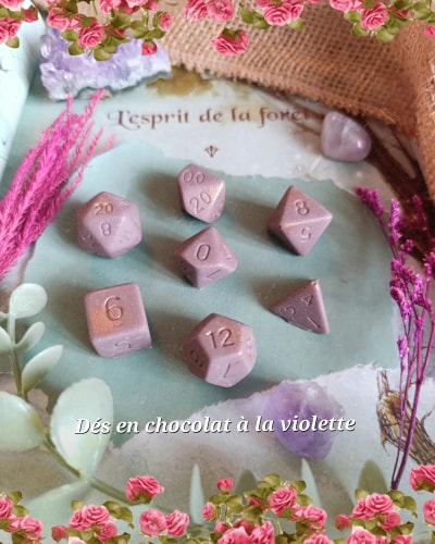 Dés en chocolat rose poudré décorés de chiffres, disposés sur un tissu pastel, entourés de fleurs roses, de feuillage et d’accessoires violets.