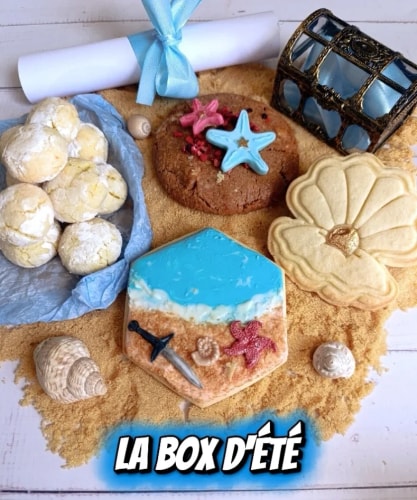 Une composition de pâtisseries artisanales, tablettes de chocolat décorées, dés de jeu de rôle mauves et enveloppes kraft sur une surface claire, agrémentée de fleurs séchées.