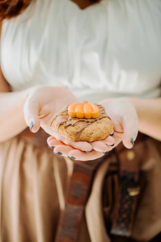 Une composition de pâtisseries artisanales, tablettes de chocolat décorées, dés de jeu de rôle mauves et enveloppes kraft sur une surface claire, agrémentée de fleurs séchées.