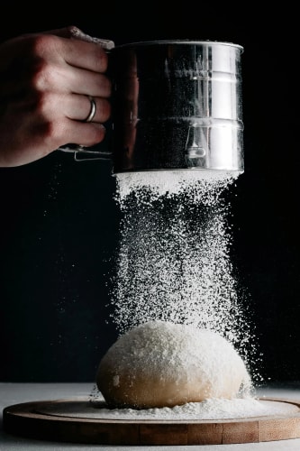 Une main tenant un tamis en métal saupoudre de la farine sur une boule de pâte à pain posée sur une planche en bois, avec un fond noir.