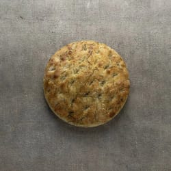 Pain rond grillé aux herbes posé sur une surface grise.