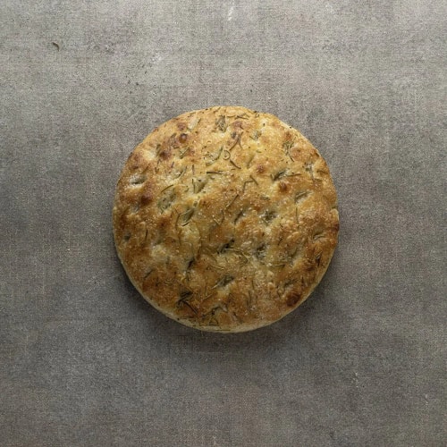 Pain rond grillé aux herbes posé sur une surface grise.