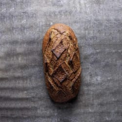 Une miche de pain artisanal brun avec une croûte gravée en motifs géométriques, posée sur une surface grise rugueuse.