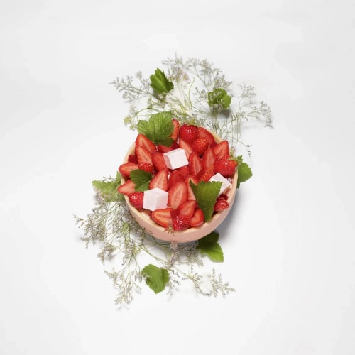 Bol rond contenant des fraises coupées en tranches, des fraises entières, des cubes blancs, décoré avec des feuilles vertes et des fleurs blanches sur fond blanc.