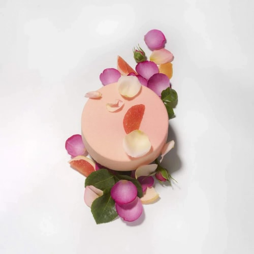 Gâteau rond rose clair décoré avec des pétales de fleurs roses et blanches ainsi que des tranches d'agrumes sur une surface blanche.