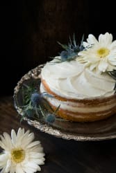 Gâteau à deux couches avec glaçage blanc, décoré de fleurs blanches et de chardons bleus, posé sur un plateau argenté, avec une fleur blanche posée à côté sur une surface en bois foncé.