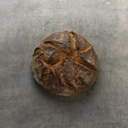 Pain artisanal rond avec une croûte croustillante et des motifs dorés sur le dessus, posé sur une surface grise.