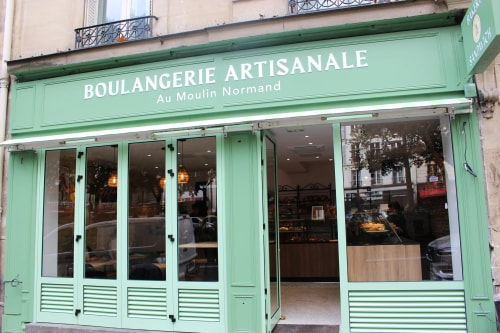 Devanture d'une boulangerie avec plusieurs baguettes de pain exposées dans la vitrine en bois, un homme en train de manipuler du pain à l'intérieur et une porte ouverte à droite où une personne est visible.