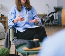 Une personne assise dans un salon, vêtue d'une chemise bleue, présentant quelque chose, pour représenter le consultant Keystone Recrutement à votre écoute.