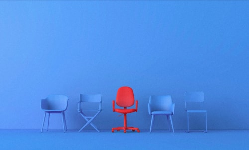 Une pièce avec un mur bleu et cinq chaises. Une chaise de bureau rouge est au centre, entourée de quatre chaises aux teintes de bleu.