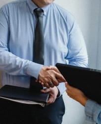 Un homme en chemise bleue et cravate noire serre la main d'une autre personne hors champ, tenant une tablette et des documents, pour représenter l'accompagnement sur mesure par Keystone Recrutement.
