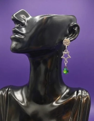 Un buste noir élégant portant une boucle d'oreille fantaisie composée d'un disque texturé, d'une étoile argentée et d'une perle en forme de goutte verte, sur fond violet.