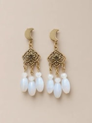 Paire de boucles d'oreilles dorées, composées d'un croissant de lune, d'un élément floral ajouré et de trois pampilles en pierre blanche ovale, sur fond beige.
