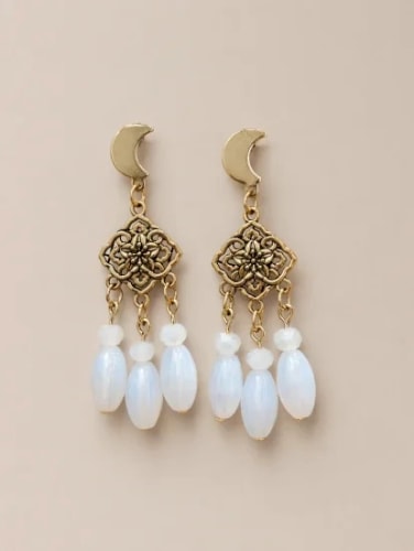 Paire de boucles d'oreilles dorées, composées d'un croissant de lune, d'un élément floral ajouré et de trois pampilles en pierre blanche ovale, sur fond beige.