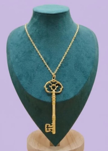 Collier doré avec un pendentif en forme de clé ancienne, présenté sur un buste de bijoutier en velours vert foncé, devant un fond violet clair.