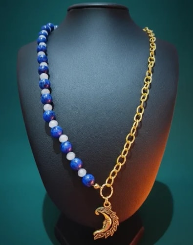 Collier asymétrique sur un buste noir : une moitié est faite de perles bleues et blanches, l'autre d'une chaîne dorée avec un pendentif en forme de lune sculptée.