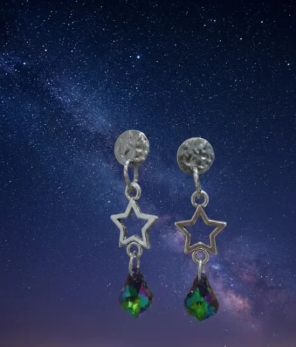 Paire de boucles d'oreilles pendantes avec un disque texturé, une étoile ajourée et une perle en cristal irisé, suspendues sur un fond de ciel étoilé et de galaxie.