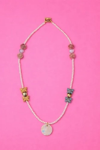 Collier de perles blanches agrémenté de perles décoratives en forme de nœuds dorés et bleus, de perles rondes et d'un pendentif circulaire central sur fond rose.