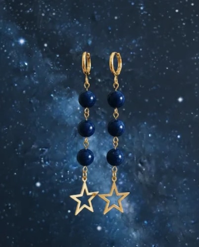 Paire de boucles d'oreilles dorées pendantes composées de trois perles bleu marine et d'une étoile ajourée à l'extrémité, présentées sur un fond représentant une galaxie étoilée.