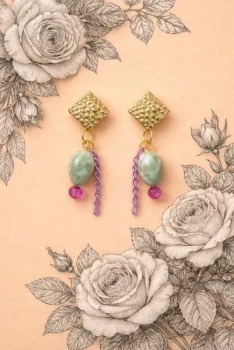 Paire de boucles d'oreilles dorées en forme de losange avec pampilles, perles vert pâle, petites perles fuchsia et chaînettes mauves sur fond rose illustré de roses dessinées au trait.