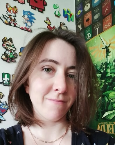 Selfie d'une jeune femme aux cheveux châtains, souriant devant un mur décoré de figurines pixelisées de jeux vidéo et d'une affiche Zelda.