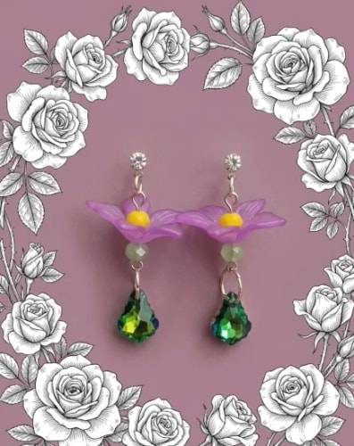 Paire de boucles d'oreilles pendantes avec une fleur violette, une petite perle et une goutte en cristal vert, sur un fond rose entouré d'un cadre de roses dessinées en noir et blanc.
