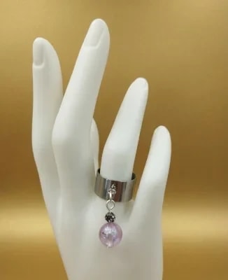 Bague en métal argenté large avec une breloque pendante composée d'une perle rose et d'un cristal, présentée sur un présentoir en forme de main blanche, sur fond doré.
