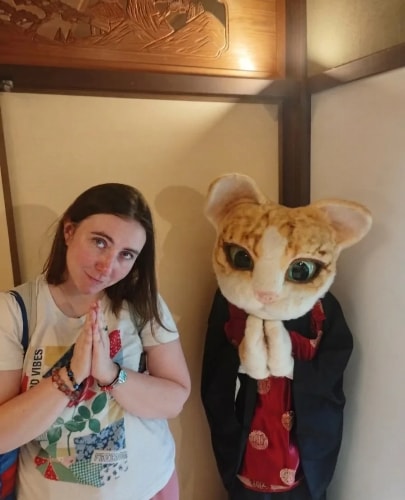 Une jeune femme et une mascotte de chat anthropomorphe en kimono sont côte à côte, les mains jointes en signe de prière devant un mur intérieur.