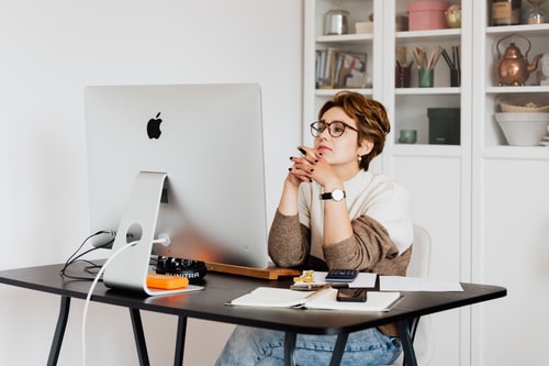 Femme assise à un bureau, regardant un grand écran d'ordinateur, le menton soutenu par ses mains. Elle porte des lunettes et un pull beige, avec des notes et un calculateur sur le bureau. En arrière-plan, une étagère avec divers objets.